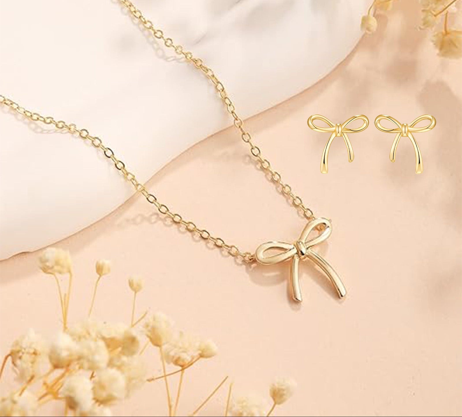 Gold bow-shaped necklace and earrings on a soft beige background with dried flowers.