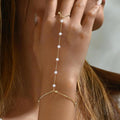 Gold hand chain with pearls worn on a hand against a neutral background