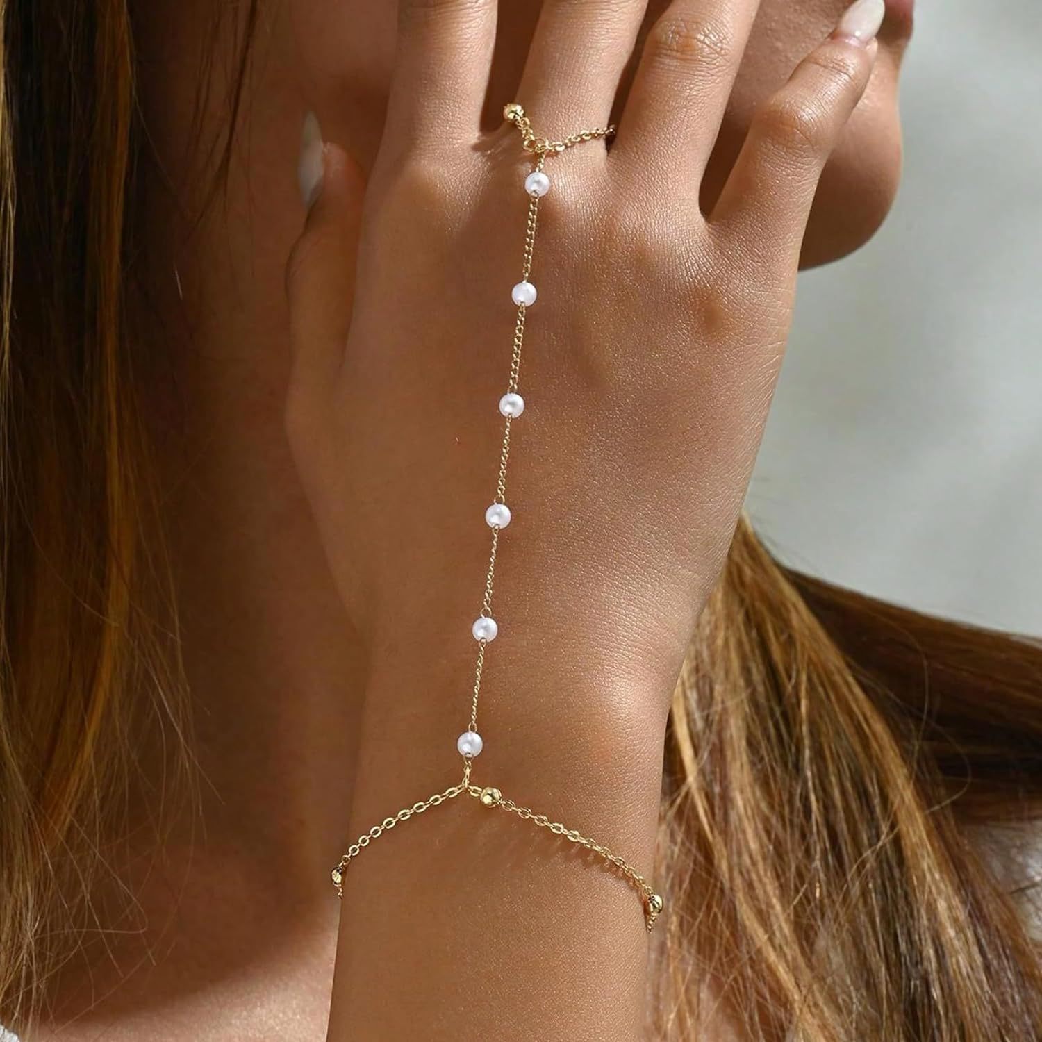Gold hand chain with pearls worn on a hand against a neutral background
