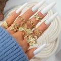 Hand wearing gold butterfly and floral rings with white nail polish on a textured surface