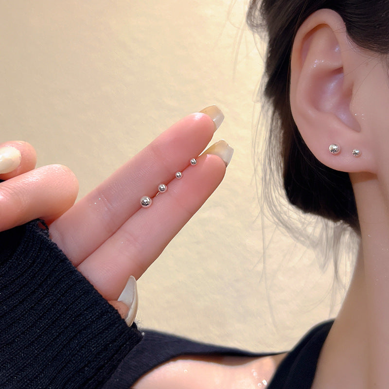 Close-up of an ear with silver stud earrings and a hand holding more earrings against a neutral background.