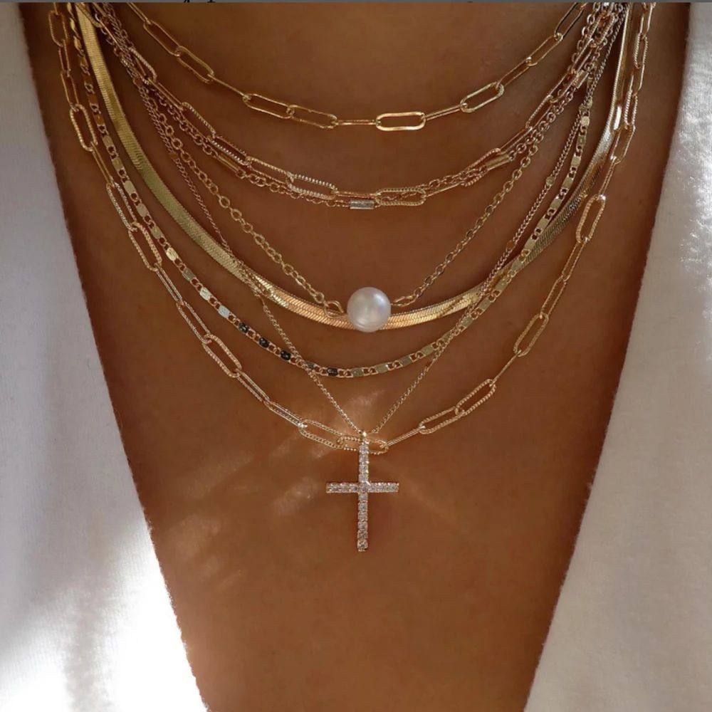 Multiple gold chains with a cross pendant and pearl on a person's neck against a neutral background